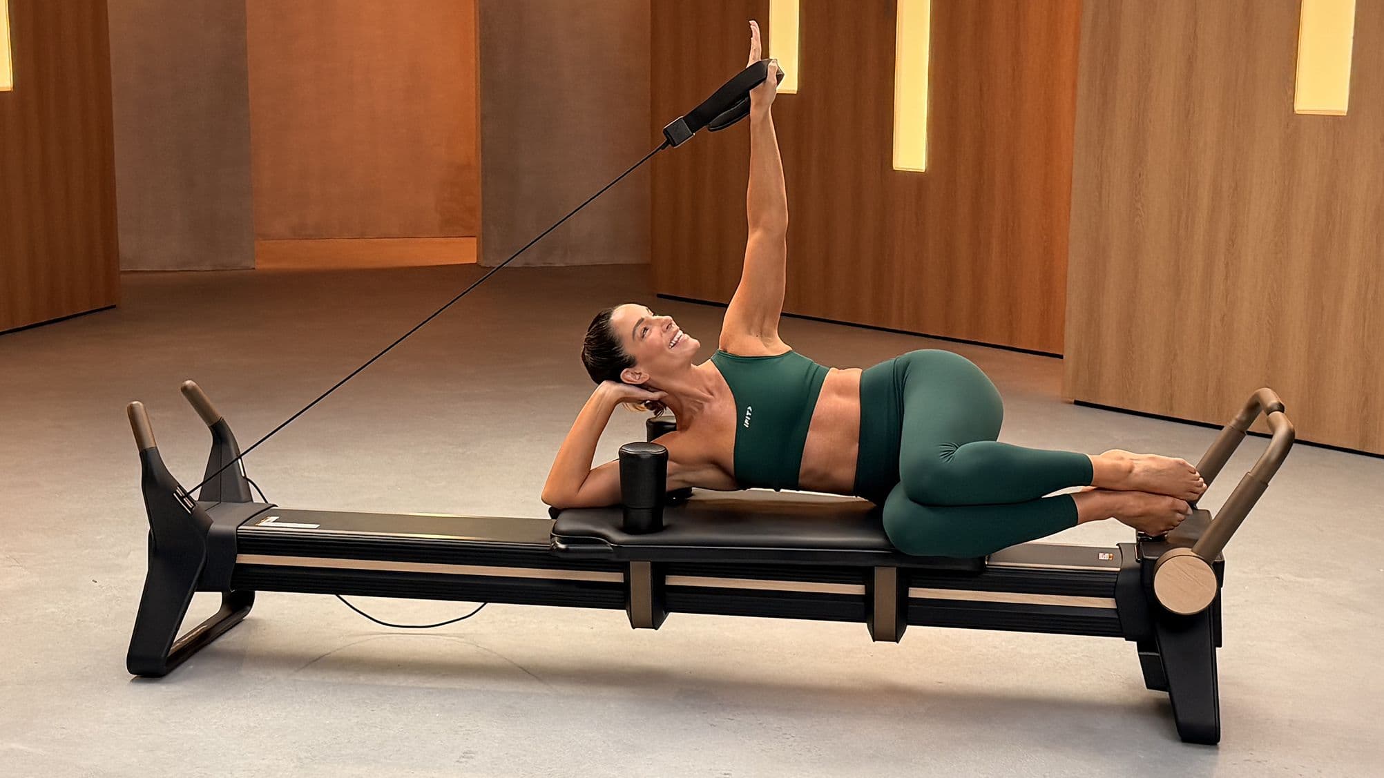 iFIT Trainer Jeni DelPozo performing a side-lying reformer Pilates exercise on the NordicTrack Ultra 1 Reformer, arm reaching overhead.