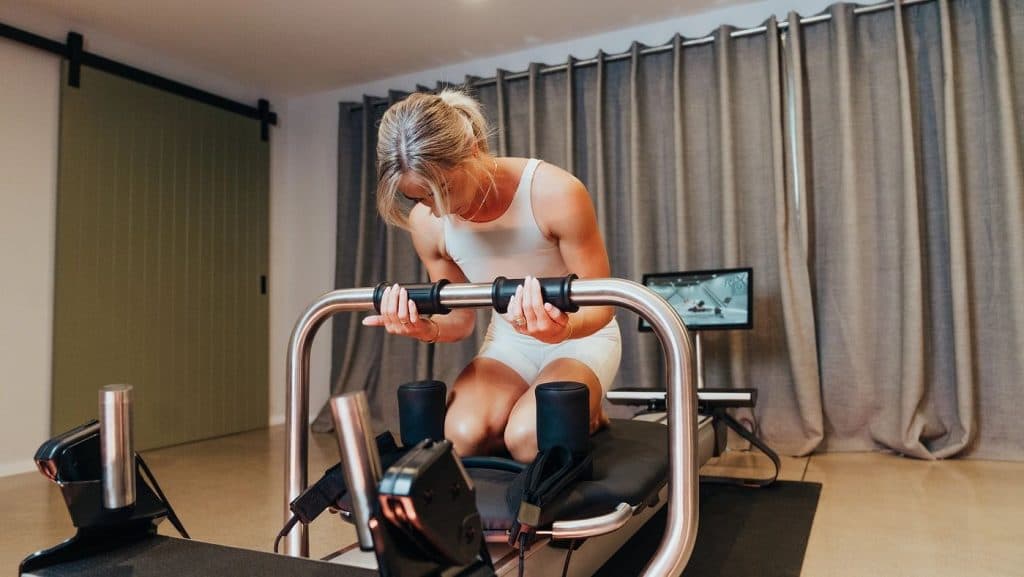 Individual performing controlled arm work while kneeling on a connected Pilates reformer in a home setting.