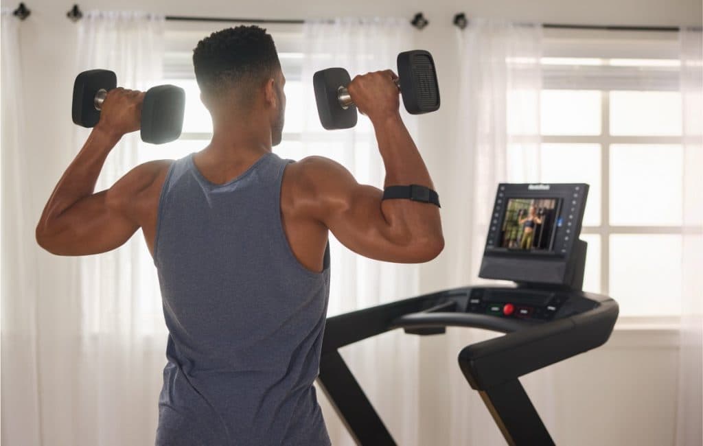 Individual performing a dumbbell shoulder press while following a guided iFIT workout on a NordicTrack treadmill screen at home.