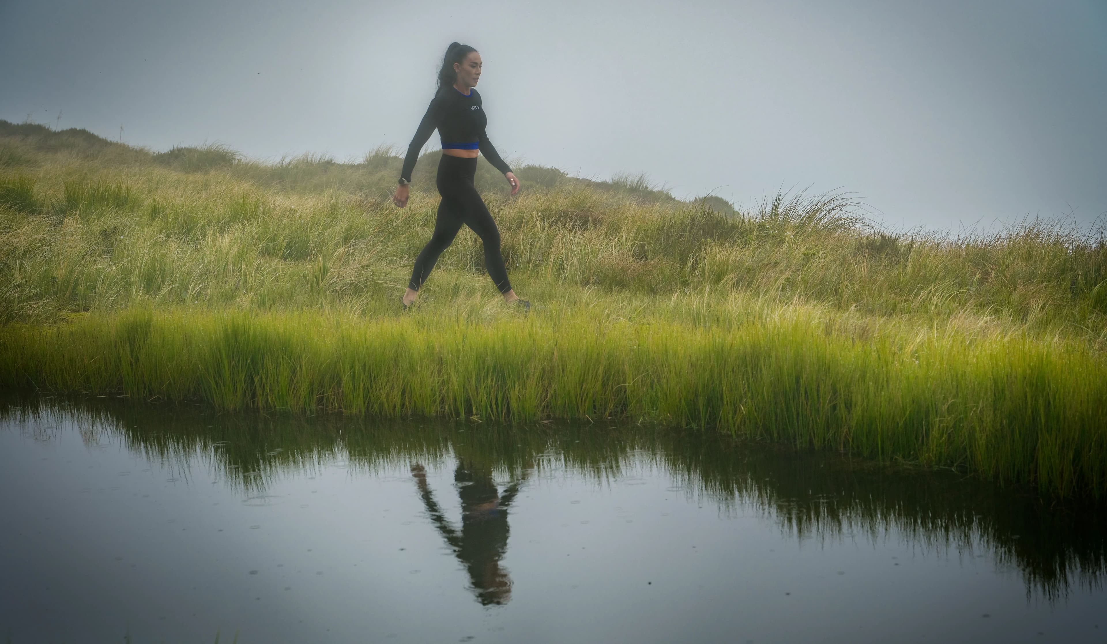 iFIT trainer Hannah Eden walking along a grassy hillside trail beside a reflective pool in New Zealand during an iFIT outdoor hiking workout