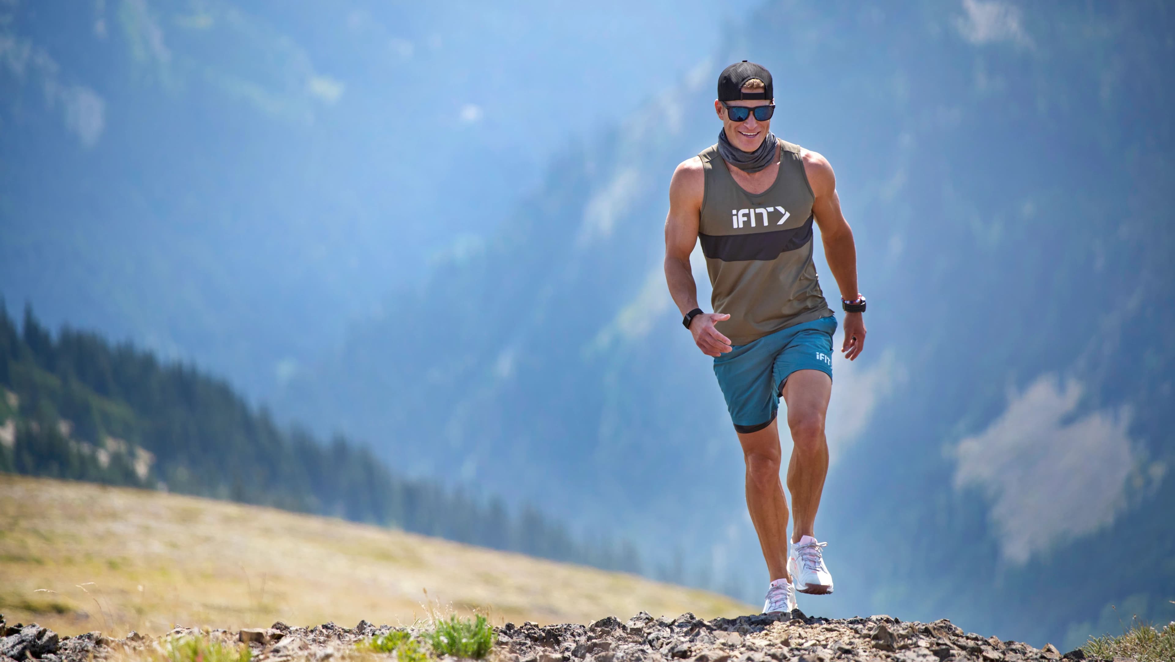 iFIT Trainer John Peel walking up a rocky alpine trail at Olympic National Park with mountain peaks and forest in the background