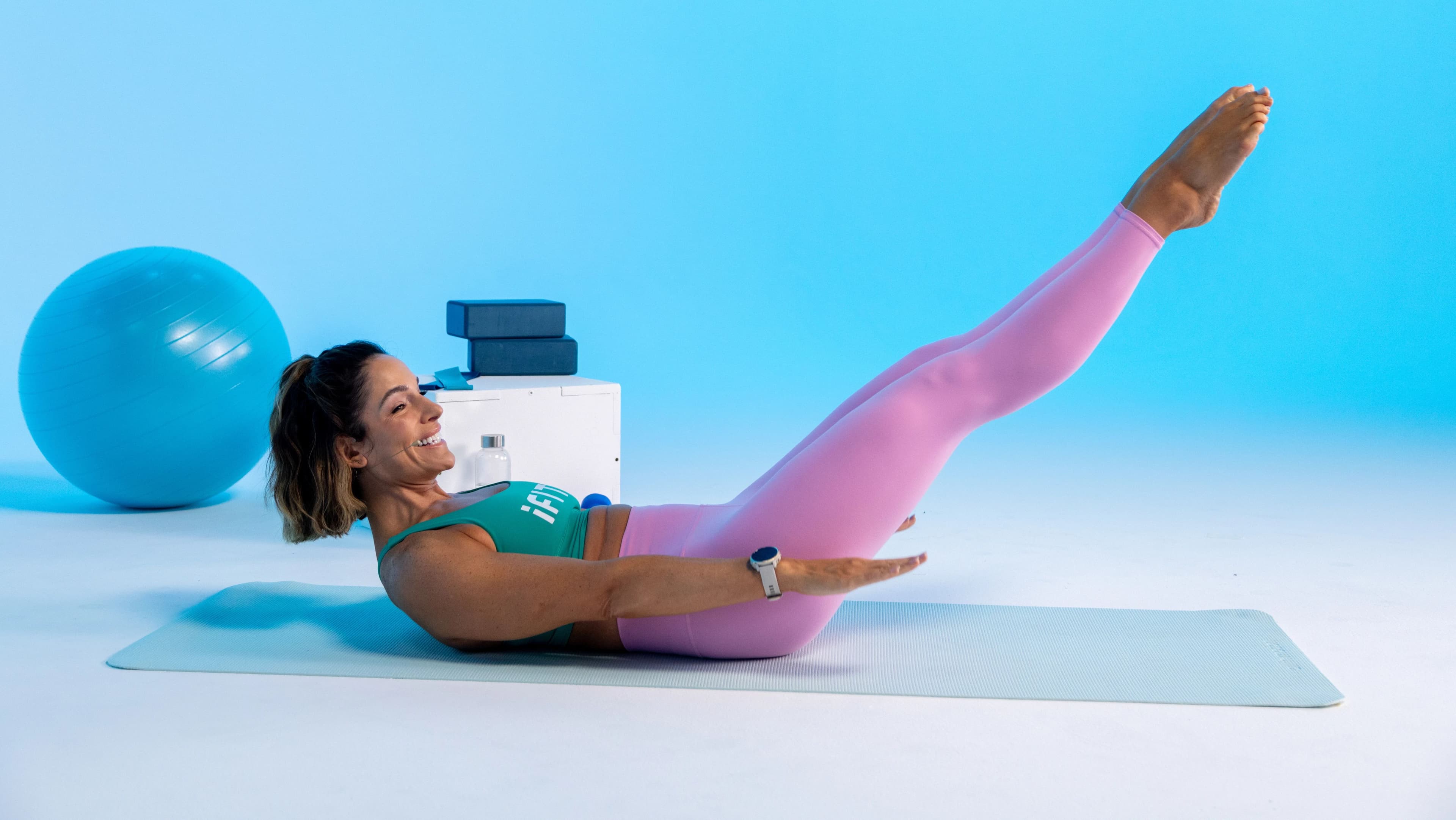iFIT trainer Jeni DelPozo performing a Pilates leg raise on a mat with a stability ball and yoga blocks in the background