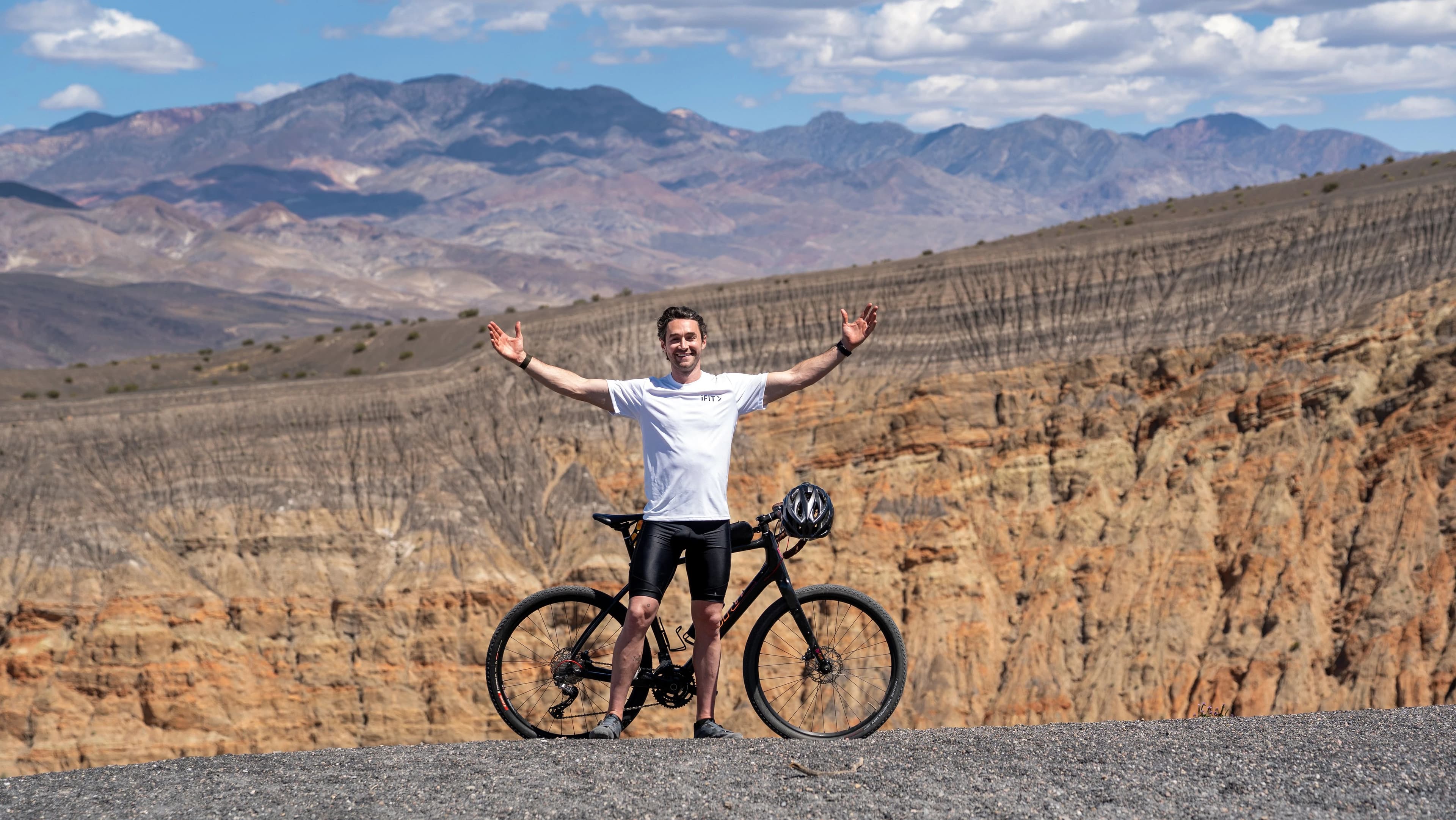iFIT Trainer Casey Gilbert standing with arms raised beside a mountain bike at Death Valley National Park with layered canyon walls behind him