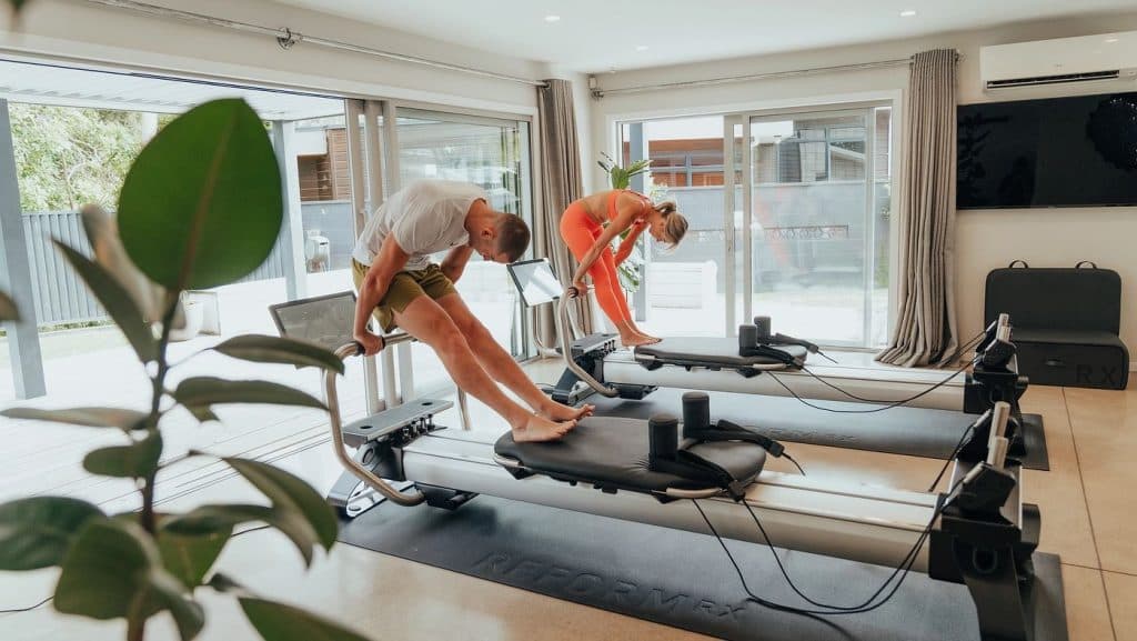 Two individuals performing reformer Pilates exercises in a bright home environment with connected screens.